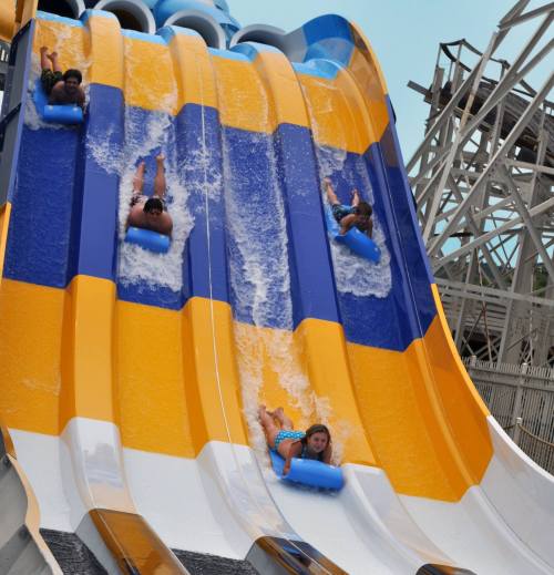 Four people slide down a colorful water slide, splashing into the pool below on a sunny day.