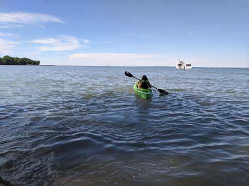 A person paddles a green kayak on calm water, with a boat in the distance and a clear blue sky above.