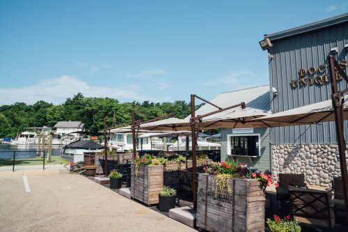A waterfront restaurant with outdoor seating, surrounded by boats and greenery under a clear blue sky.
