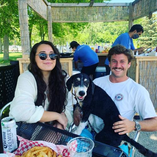 A woman and a man sit at a table with a dog, enjoying food and drinks in a sunny outdoor setting.