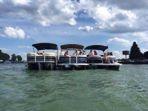 Four pontoon boats anchored together on a lake, with people relaxing under canopies on a sunny day.