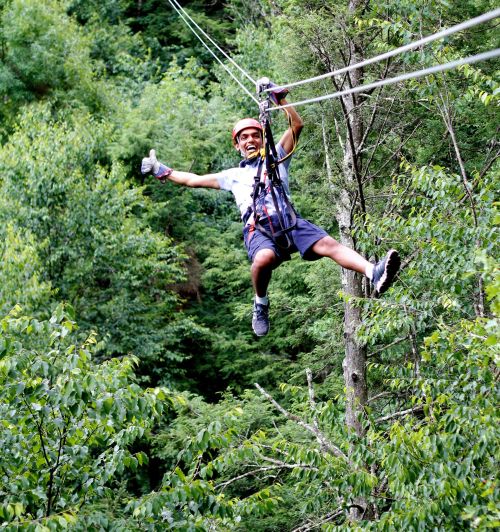 A person in a helmet zip-lining through a lush green forest, smiling and giving a thumbs up.