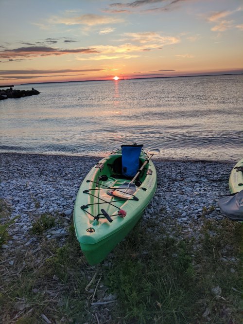 A green kayak on a rocky shore at sunset, with calm water reflecting the colorful sky.