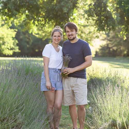 A couple stands together in a sunny field, smiling and holding lavender flowers. Green trees are in the background.