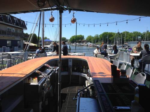 A waterfront bar with a wooden counter, patrons enjoying drinks, and boats docked in the background on a sunny day.