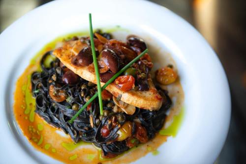 A plate of black pasta topped with grilled fish, olives, cherry tomatoes, and garnished with chives.