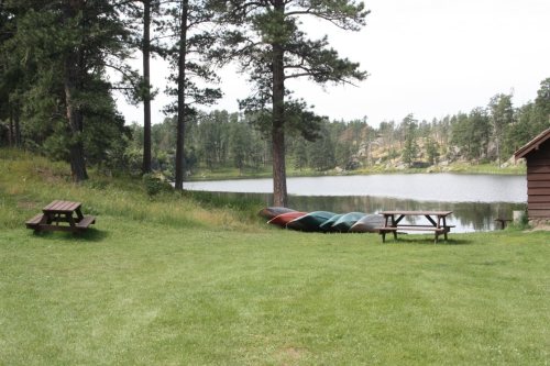 A serene lakeside scene with canoes by the water, a picnic table on the grass, and trees in the background.
