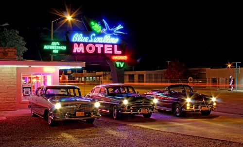 Three classic cars parked in front of the Blue Swallow Motel, illuminated by a vibrant neon sign at night.