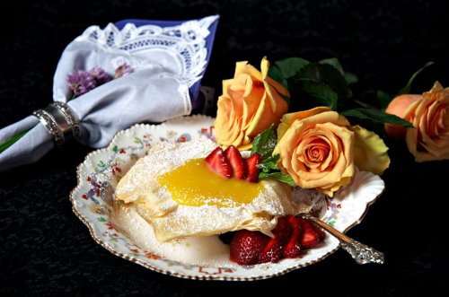 A delicate dessert on a floral plate, topped with lemon sauce and strawberries, accompanied by roses and a lace napkin.