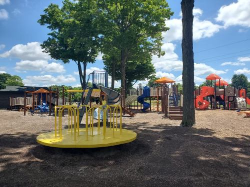 A colorful playground with slides, climbing structures, and a yellow merry-go-round, surrounded by trees and blue skies.