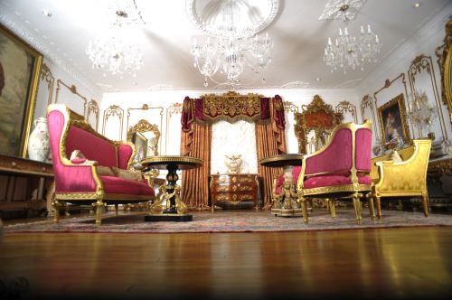 Luxurious room with ornate furniture, chandeliers, and rich decor, featuring pink and gold seating and elegant artwork.