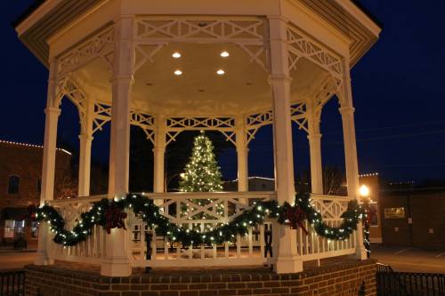 A decorated gazebo at night, featuring a Christmas tree and festive lights, surrounded by greenery and ribbons.