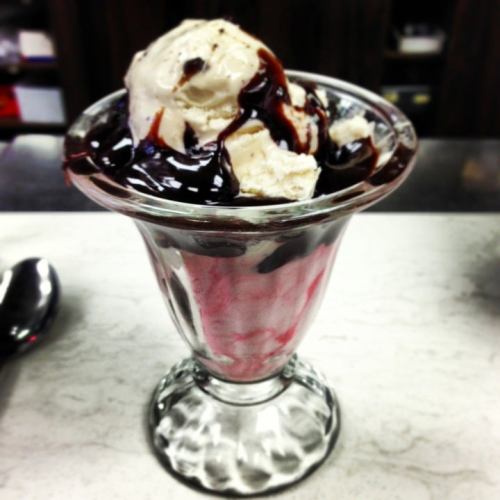 A glass dessert dish filled with layers of pink ice cream, topped with chocolate syrup and a scoop of vanilla ice cream.