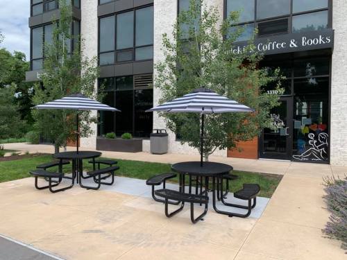Two black picnic tables with striped umbrellas outside a coffee shop and bookstore, surrounded by greenery.