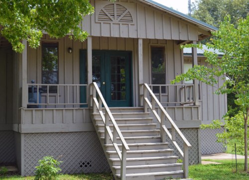 A light-colored house with a front porch and stairs, surrounded by greenery and trees.
