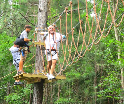 Two people on a wooden platform in a forest, navigating a rope course with trees in the background.