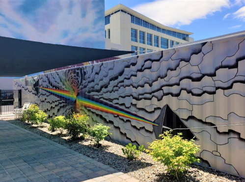 Colorful mural featuring a rainbow and abstract patterns on a wall, with greenery and a modern building in the background.