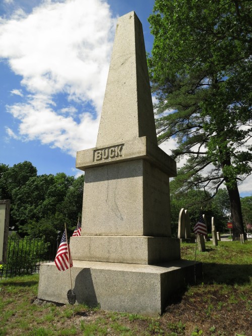 A tall granite obelisk monument with the name "BUCK" engraved, surrounded by trees and small American flags.