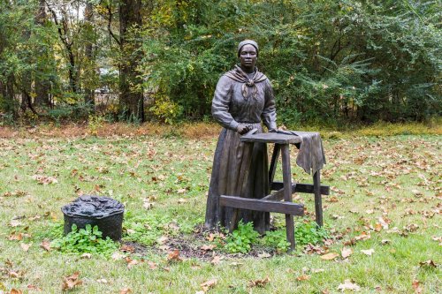 A bronze statue of a woman in period clothing, standing by a washboard in a grassy area surrounded by trees.
