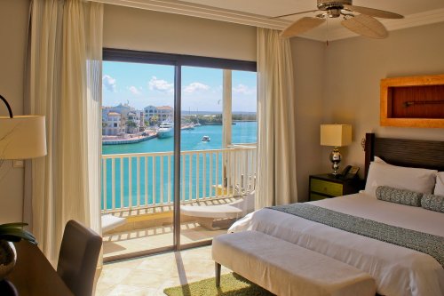 A bright hotel room with a bed, overlooking a balcony and a scenic view of the water and buildings.