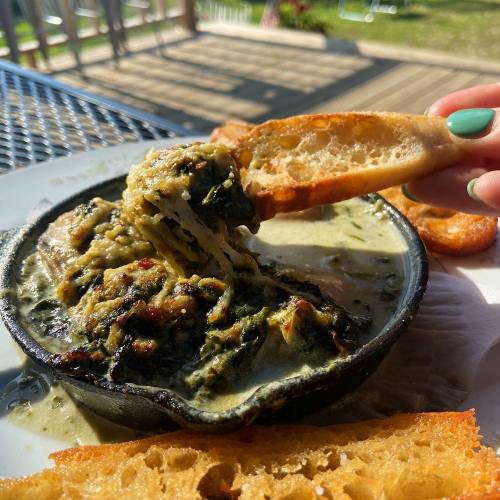 A creamy spinach dish in a black bowl, served with toasted bread on a sunny outdoor table.
