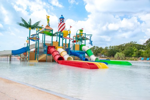 Colorful water slides and play structures in a pool area, surrounded by palm trees and a blue sky with clouds.