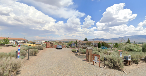 A gravel road leads through a desert area with sparse vegetation, buildings, and mountains under a cloudy sky.