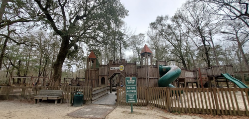 A wooden playground with slides and towers, surrounded by trees and a sign stating "No Pets Allowed."