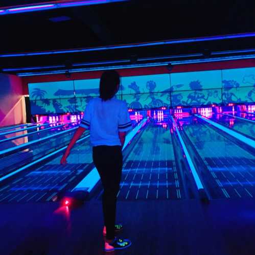 A person stands at a bowling alley, illuminated by colorful lights, with bowling lanes stretching ahead.