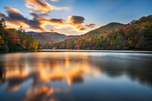 Serene lake reflecting colorful autumn trees and mountains under a vibrant sunset sky.