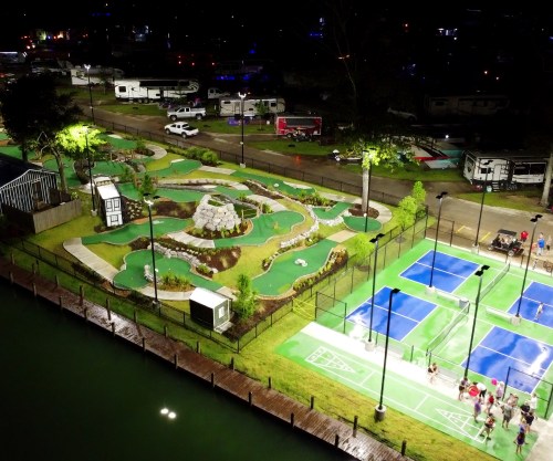 Aerial view of a recreational area at night featuring mini-golf, tennis courts, and RVs by a water body.
