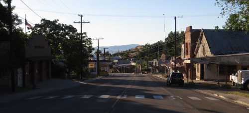 A quiet street in a small town, lined with buildings and trees, under a clear blue sky. Mountains are visible in the distance.