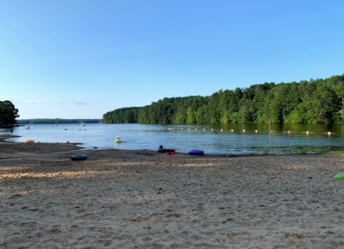 A sandy beach by a calm lake surrounded by green trees, with people enjoying the water and floating on colorful rafts.