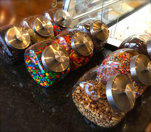 A row of clear jars filled with colorful candies and toppings, featuring metallic lids, on a dark countertop.