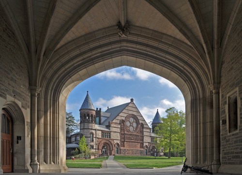 A historic building with intricate architecture framed by an arched stone entrance, surrounded by greenery and blue skies.