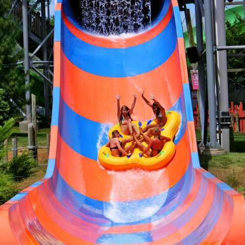 A group of children joyfully riding a yellow inflatable raft down a colorful water slide, splashing water around.