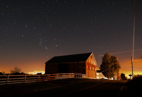 A barn under a starry night sky, with a road and trees illuminated by distant lights.