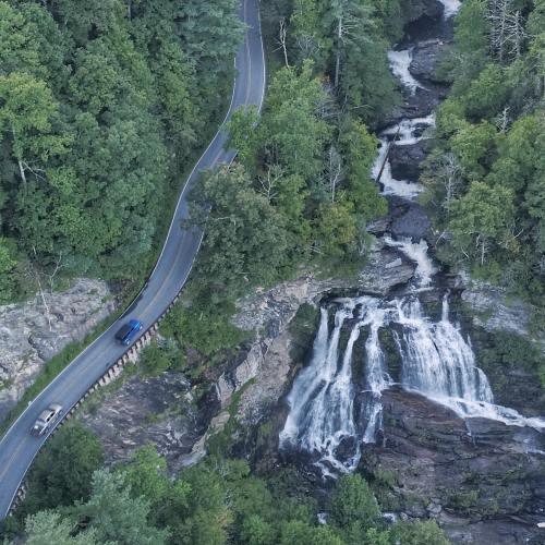 Aerial view of a winding road beside a waterfall, surrounded by lush green trees.