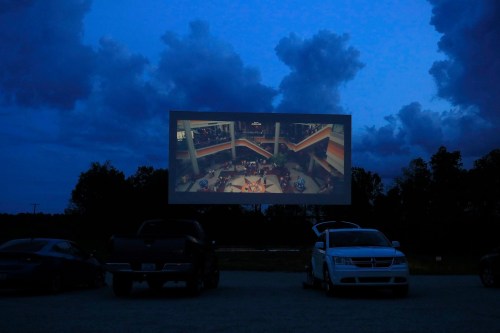 A drive-in movie screen displays a film against a twilight sky with clouds, surrounded by parked cars.