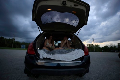 Two people sitting in the back of a car with a blanket, under a cloudy sky in a parking area.