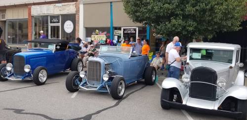 Three classic cars in vibrant colors parked on a street, with a crowd of people enjoying a car show in the background.