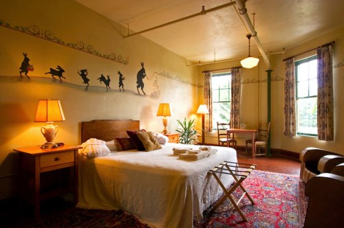 Cozy bedroom with a bed, two lamps, a rug, and whimsical wall art of silhouettes dancing along the wall.