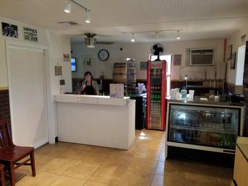 A cozy café interior with a counter, a drink fridge, and a woman standing behind the counter.