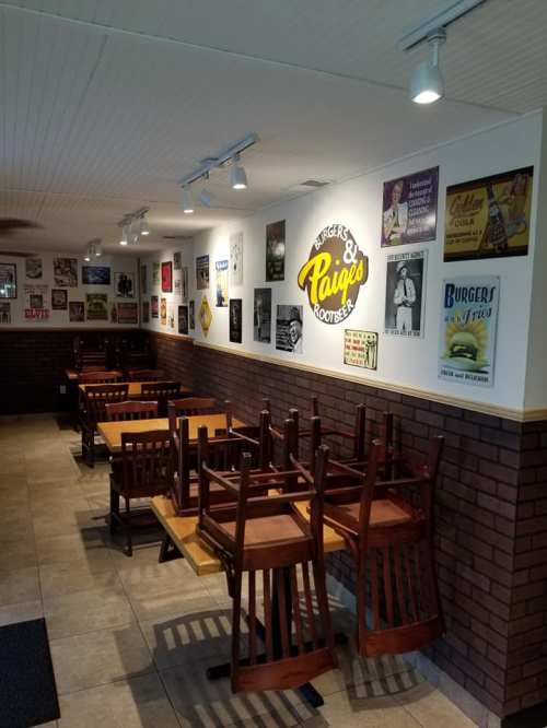 A cozy restaurant interior with stacked chairs, a sign for "Burgers & Paige's Root Beer," and vintage posters on the walls.