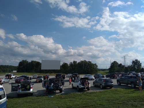 A drive-in movie theater with cars parked, people sitting outside, and a large blank screen under a cloudy sky.