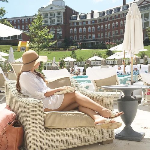A woman in a white dress and hat relaxes on a lounge chair by a pool, surrounded by umbrellas and greenery.