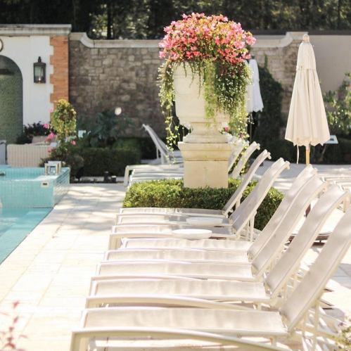A serene poolside scene with lounge chairs, potted flowers, and a stone wall in a sunny outdoor setting.