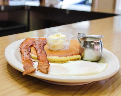A plate of breakfast featuring pancakes, bacon, eggs, butter, and syrup on a wooden table.