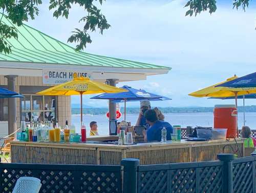 A beachside bar with colorful umbrellas, patrons enjoying drinks, and a scenic view of the water in the background.