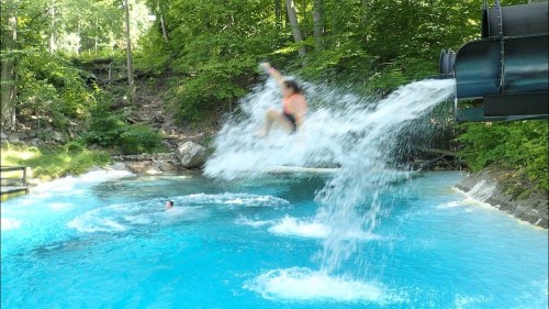 A person jumps from a water slide into a blue pool, creating a splash, while another person swims nearby.